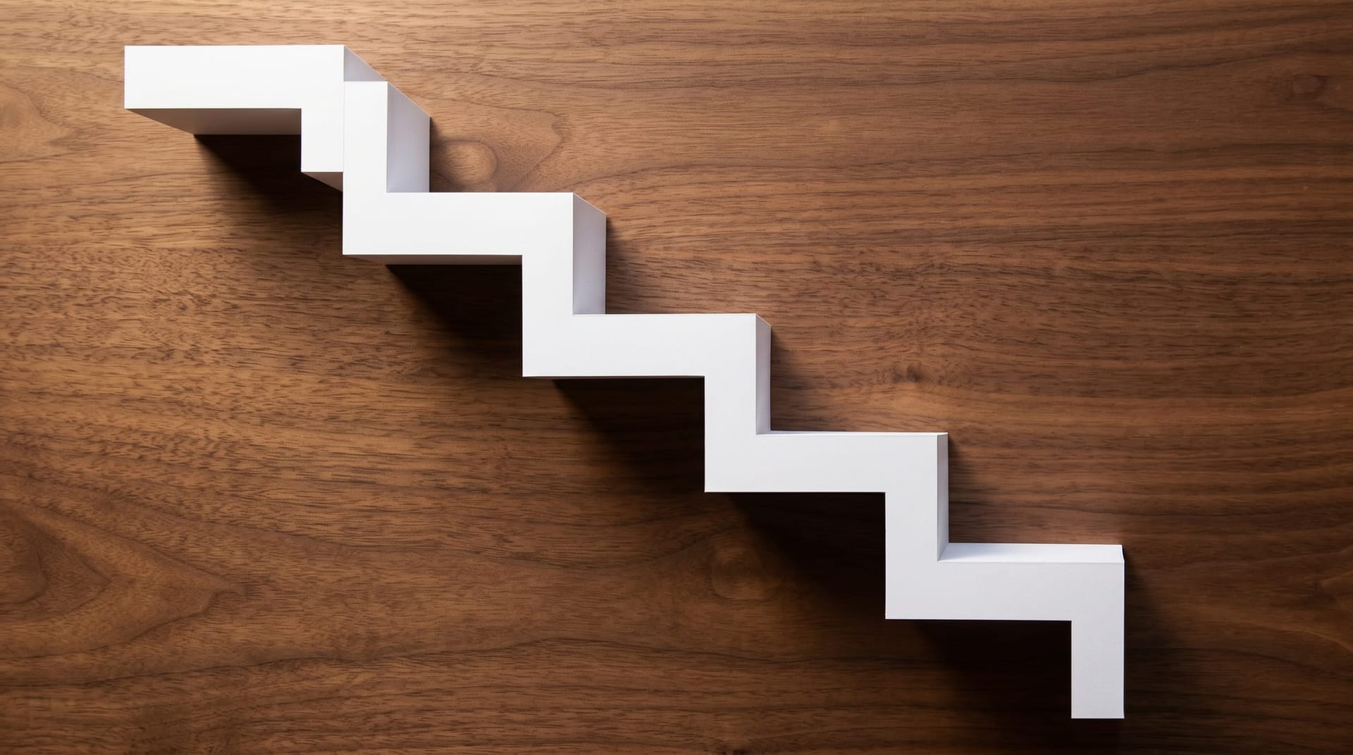 An overhead flat-lay of a precisely cut white paper staircase descending across a deep walnut surface, evoking the cost cliff that just dropped video AI pricing 80–90 percent.