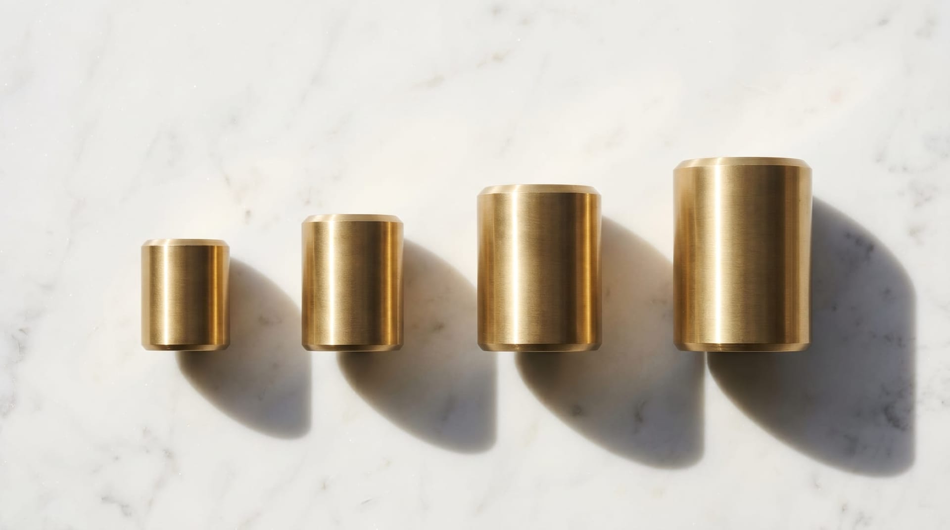 Four identical brushed-brass weights of increasing height arranged in a row on a marble surface, depicting the four capability axes that improve when world models ship inside the agent layer.